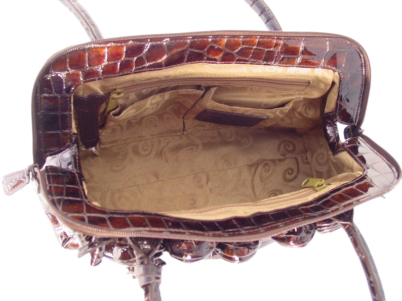 Pu&ograve; includere: Una borsa a mano satchel marrone in pelle con stampa coccodrilo con chiusura a zip superiore e un interno in tessuto marrone. La borsa a mano ha un manico superiore e una tracolla rimovibile.