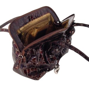 Pu&ograve; includere: Una borsa a mano in pelle marrone con stampa coccodrilo e un ciondolo a forma di cuore. La borsa ha una chiusura con cerniera superiore e due manici.