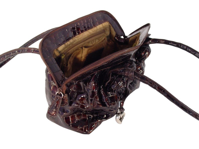 Pu&ograve; includere: Una borsa a mano in pelle marrone con stampa coccodrilo e un ciondolo a forma di cuore. La borsa ha una chiusura con cerniera superiore e due manici.