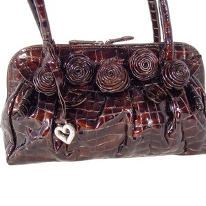 Pu&ograve; includere: Una borsa a mano in pelle marrone con effetto coccodrilo, con un ciondolo a forma di cuore e decorazioni floreali.