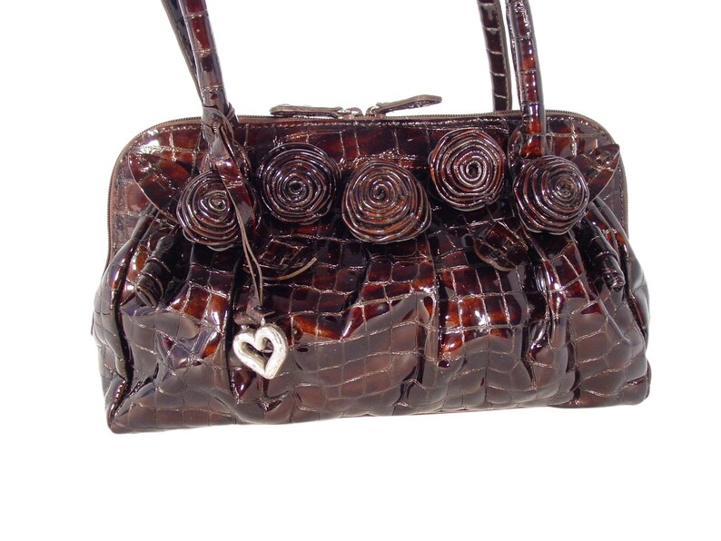 Pu&ograve; includere: Una borsa a mano in pelle marrone con effetto coccodrilo, con un ciondolo a forma di cuore e decorazioni floreali.