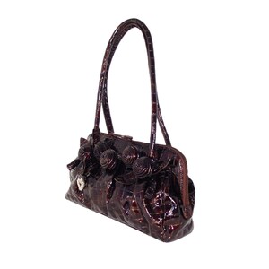 Pu&ograve; includere: Una borsa a mano in pelle marrone con effetto coccodrilo, con un design floreale decorativo e un ciondolo a forma di cuore argentato.