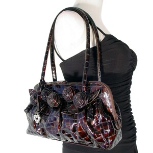 Pu&ograve; includere: Una borsa a mano marrone in pelle di coccodrilo con un ciondolo a forma di cuore e dettagli decorativi a forma di rosa.