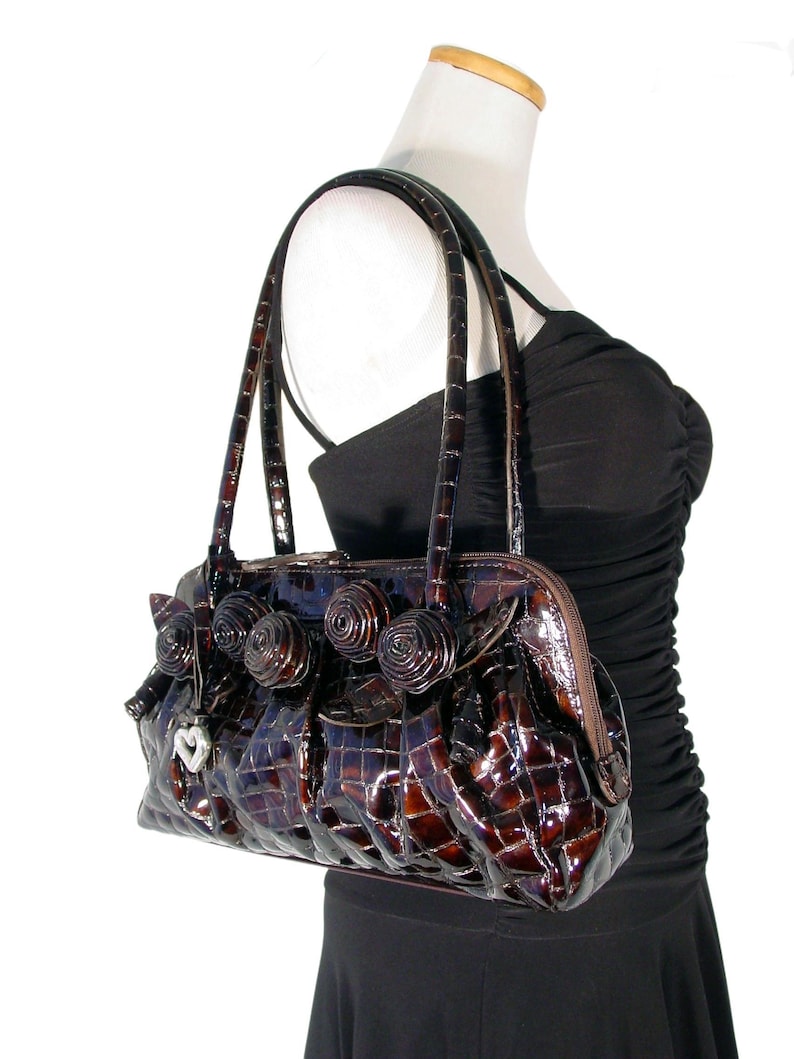 Pu&ograve; includere: Una borsa a mano marrone in pelle di coccodrilo con un ciondolo a forma di cuore e dettagli decorativi a forma di rosa.