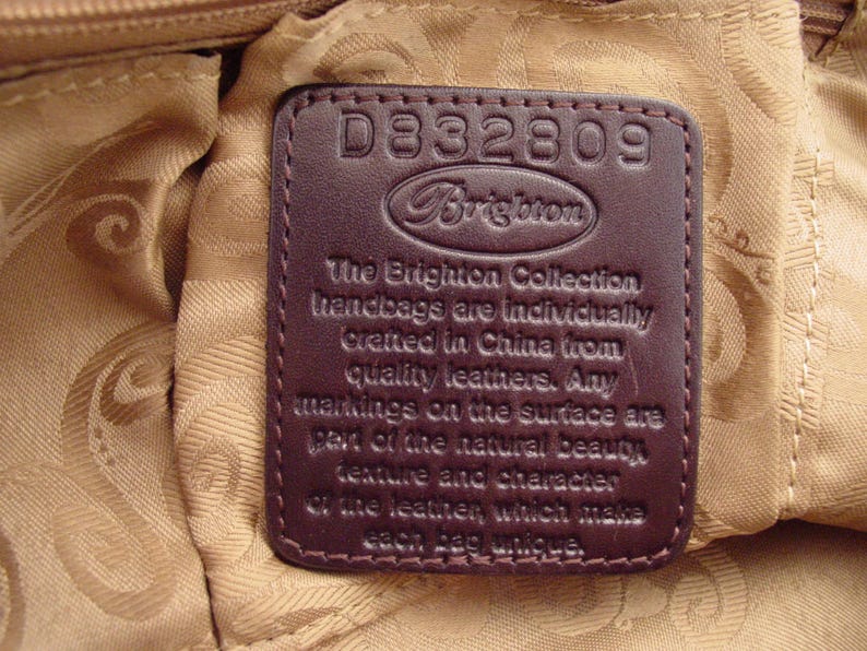 Pu&ograve; includere: Un'etichetta per borsa a mano in pelle marrone con il testo "D332809 Brighton The Brighton Collection handbags are individually created in China from quality leathers. Any markings on the surface are part of the natural beauty, texture and character of the leather which makes each bag unique."