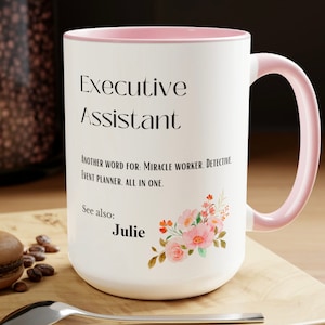 Könnte beinhalten: Eine weiße Keramik-Kaffeetasse mit rosafarbenem Rand und Henkel. Die Tasse hat einen schwarzen Text, der "Executive Assistant" und "Another word for: Miracle Worker, Detective, Event Planner. All in one." lautet. Die Tasse hat auch ein Blumenmuster mit rosa und orangefarbenen Blumen und den Namen "Julie" in schwarzer Schrift.