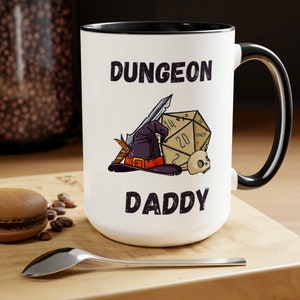 May include: White ceramic mug with a black rim and handle. The mug has a black and white illustration of a wizard's hat, a sword, and a 20-sided die. The text "Dungeon Daddy" is printed in black on the mug.