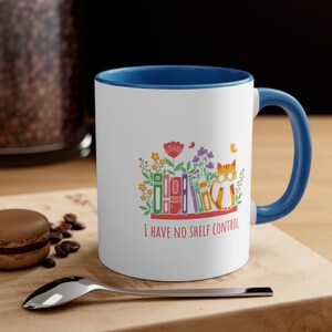 Könnte beinhalten: Eine weiße Keramiktasse mit blauem Rand und Henkel. Die Tasse zeigt eine bunte Illustration einer Katze mit Brille, die auf einem Stapel Bücher mit Blumen drumherum sitzt. Der Text "I have no shelf control" ist auf die Tasse gedruckt.