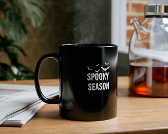 May include: Black ceramic mug with the text "SPOOKY SEASON" in white, and bat silhouettes. The mug is on a wooden surface, with steam rising from it. A glass carafe is in the background.