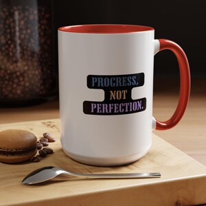 May include: White ceramic mug with a red interior and handle. The mug features the text "PROGRESS, NOT PERFECTION" in a black, blue, and yellow design. A spoon and a macaron are on a wooden surface.