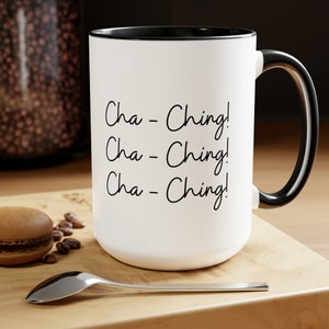Könnte beinhalten: Weiße Keramik-Kaffeetasse mit schwarzem Rand und Henkel. Die Tasse trägt den Text "Cha-Ching! Cha-Ching! Cha-Ching!" in schwarzer Kursivschrift.