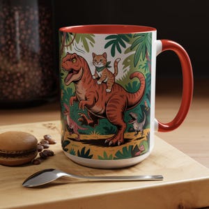 May include: White ceramic mug with a red interior and handle. The mug features a colorful illustration of a cat riding a dinosaur in a jungle setting. Other dinosaurs and foliage are also depicted.