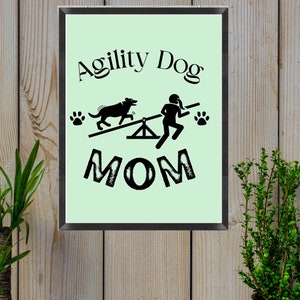 May include: A black and white graphic design with the text "Agility Dog Mom" on a light green background. The graphic shows a dog jumping over a seesaw and a person running next to it.