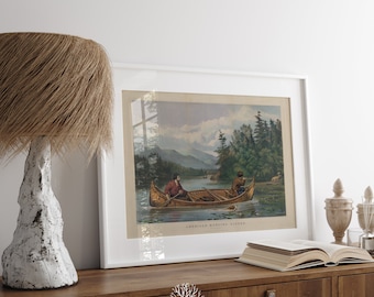 May include: Framed vintage print titled "American Hunting Scene" depicting two men in a canoe on a lake, surrounded by trees and mountains. The print is in a white frame, displayed on a wooden cabinet with a lamp and decorative items.