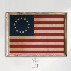 Puede incluir: Una impresión enmarcada de la bandera de Betsy Ross, la primera bandera estadounidense, con 13 estrellas en un círculo sobre un campo azul y 13 rayas rojas y blancas alternas. La bandera está en un marco de madera rústico.