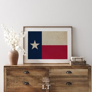 Puede incluir: Una impresión enmarcada de la bandera del estado de Texas, con un fondo azul, una estrella blanca y una franja horizontal roja. La bandera se muestra en una cómoda de madera con un jarrón de flores blancas.