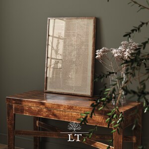 May include: A framed vintage newspaper print with text in black and white, sitting on a dark brown wooden table with a light brown tree logo and the letters "LT" underneath. A vase with white flowers and green leaves is on the table.