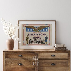 May include: Framed vintage Liberty Bond poster featuring an eagle with outstretched wings above the text "THERE IS A LIBERTY BOND IN THIS HOME." The poster has a red, white, and blue border and depicts soldiers and civilians.