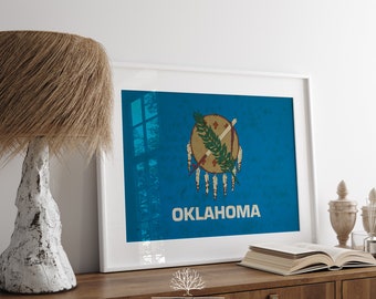 May include: A framed print of the Oklahoma state flag. The flag is blue with a white circle in the center. Inside the circle is a red and white shield with a crossed spear and bow, and a green sprig of willow. The text "OKLAHOMA" is printed in white below the circle.