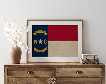 May include: A framed print of the North Carolina state flag. The flag is blue with a white star in the center and the words "MAY 20th 1775" and "APRIL 12th 1776" in gold lettering. The flag is surrounded by a white border and is displayed on a wooden dresser.