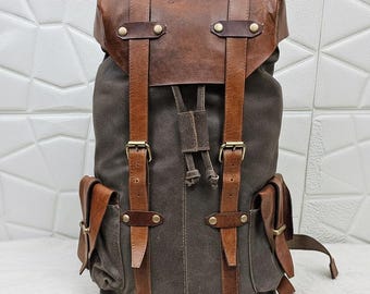 May include: A brown and gray backpack with leather straps and accents. The backpack has two side pockets and a top flap. A pair of black sunglasses and a leather tag are next to the backpack.