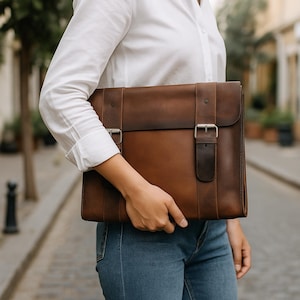 May include: A brown leather briefcase with silver buckles. The briefcase is rectangular with a flap closure and a handle. The leather has a distressed appearance. The person is wearing a white shirt and blue jeans.