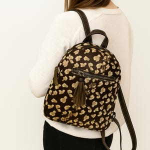 May include: A brown and tan leopard print backpack with a zipper closure and a tassel. The backpack has brown straps and a handle. The bag is made of a textured material.