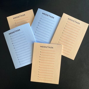 May include: Five blank note cards with the text "MEDS/TASK" printed on them. The cards are in different colors: beige, light blue, and a darker beige. Each card has a numbered list from 7 to 12, and then from 1 to 7.