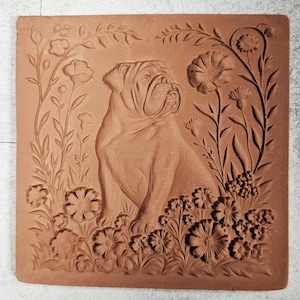May include: A square terracotta wall plaque featuring a relief carving of a bulldog sitting among various flowers and leafy vines. The natural reddish-brown clay surface displays intricate details of the dog's facial folds and floral petals.