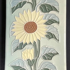 May include: A rectangular ceramic tile featuring a raised relief design of sunflowers with pale yellow petals and brown centers. The flowers are set against a light sage green background with dark green leaves and light blue accents.