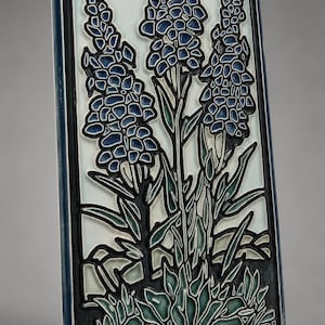 May include: A rectangular ceramic tile featuring a raised relief design of three tall blue flower stalks with green leaves against a pale off-white background. The tile has a dark blue border and a glossy finish with visible fine crackle patterns.