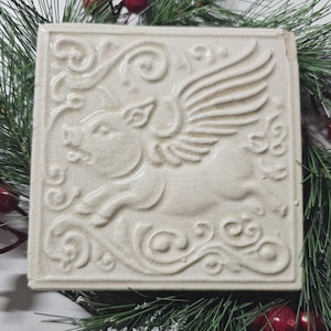 May include: A square off-white ceramic tile featuring a raised relief design of a flying pig with wings. The surface has a visible crackle glaze finish and is decorated with swirling scrollwork patterns surrounding the central pig figure.