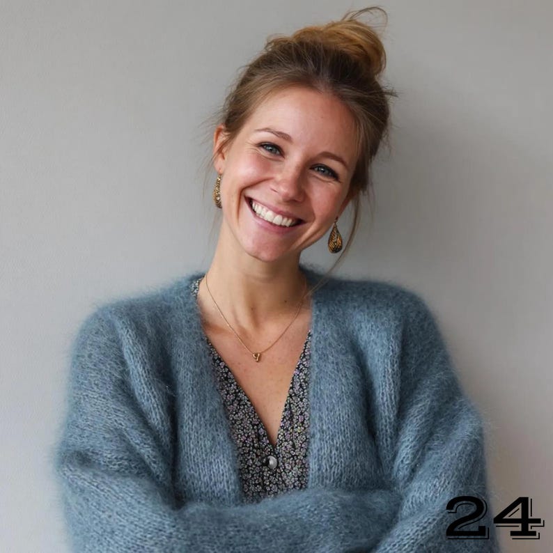 May include: A woman wearing a light blue, fluffy cardigan over a floral print dress. She is smiling, with her hair up in a bun, and wearing gold earrings and a necklace. The number "24" is in the bottom right corner.