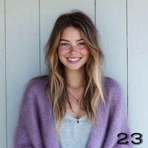 May include: A woman wearing a light purple, oversized knit cardigan over a light blue top. She has long, wavy blonde and brown hair and is smiling. A silver necklace is visible. The number "23" is in the bottom right corner.