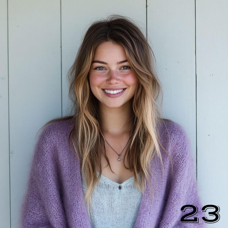 May include: A woman wearing a light purple, oversized knit cardigan over a light blue top. She has long, wavy blonde and brown hair and is smiling. A silver necklace is visible. The number "23" is in the bottom right corner.