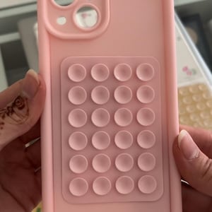 Puede incluir: Una funda de teléfono rosa claro con una sección elevada de pequeños bultos circulares. La funda tiene recortes para la cámara y el flash del teléfono. La funda está sujeta por las manos de una persona.