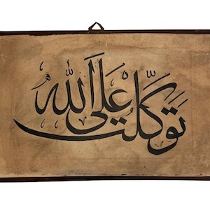 May include: Rectangular artwork featuring black Arabic calligraphy on a tan background. The artwork is framed in a dark brown border and includes a metal hanger at the top for display. The calligraphy is the focal point.