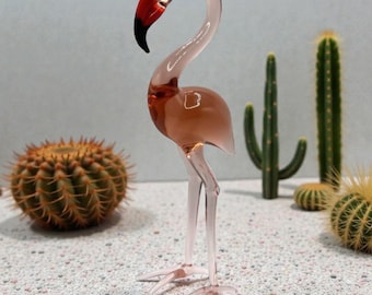 Op de afbeelding: Glazen flamingo beeldje met een roze lichaam, heldere poten en een rode en zwarte snavel. De decoratieve vogel staat op een oppervlak met kleine witte kiezelstenen, met cactussen op de achtergrond. De flamingo is een decoratief item.