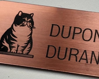 May include: A copper-colored metal name tag with a black silhouette of a cat sitting on a shelf. The text on the tag reads "DUPONT Léna DURAND Gabriel".