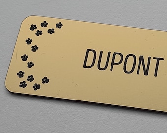 May include: A rectangular gold-colored name tag with rounded corners. The name "DUPONT Léna" is printed in black, centered on the tag. Black paw prints decorate the top left and bottom right corners.