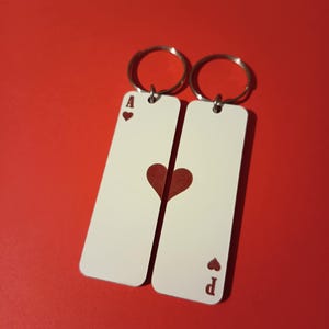 Puede incluir: Dos llaveros con aros plateados, cada uno con una etiqueta rectangular blanca. Uno muestra el As de Corazones, el otro el As de Corazones. El fondo es rojo liso.