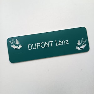 May include: A dark teal name tag with rounded corners. The name "DUPONT Léna" is in white text. Two white bird illustrations flank the name. The name tag is rectangular in shape.
