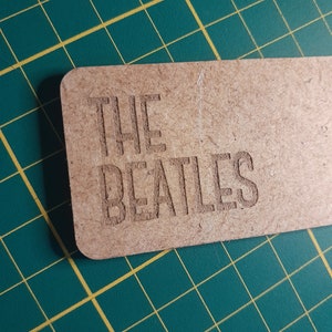 Może przedstawiać: Prostokątna, jasnobrązowa drewniana tabliczka z wygrawerowanymi słowami "THE BEATLES" pogrubioną, bezszeryfową czcionką. Tabliczka leży na zielonej macie do cięcia z żółtymi liniami siatki.