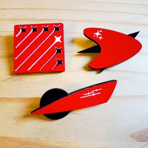 May include: Three red and black enamel pins with white starburst designs. One pin is square with diagonal lines and starbursts. Another is a boomerang shape with starbursts. The third is a red shape with a black circle.