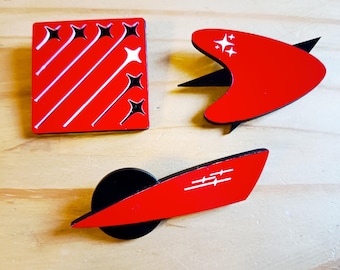 May include: Three red and black enamel pins with white starburst designs. One pin is square with diagonal lines and starbursts. Another is a boomerang shape with starbursts. The third is a red shape with a black circle.