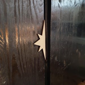 Könnte beinhalten: Ein heller, sternförmiger Schrankgriff aus Holz, montiert auf einer dunklen, glänzend schwarz gebeizten Holzoberfläche mit sichtbarer Maserung. Das Sterndesign hat scharfe, spitze Kanten und ist vertikal entlang der Schranktürfuge angebracht.
