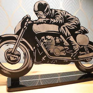 Puede incluir: Silueta negra de un motorista en una motocicleta vintage con un soporte negro. La motocicleta tiene una rueda delantera grande y una rueda trasera más pequeña.