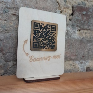 Könnte beinhalten: Ein Holzschild mit einem goldfarbenen QR-Code-Aufkleber aus Metall und dem Text "Scannez-moi" in Kursivschrift.