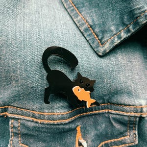 May include: A black cat brooch with an arched back and a long tail, holding a small orange fish in its mouth. The cat has pointed ears and a sleek, glossy finish, while the fish features a simple eye detail. The brooch is attached to blue denim.