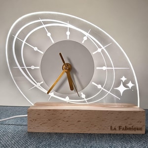 Peut inclure: Une horloge moderne et éclairée avec un cadran clair de forme ovale et une base en bois. Le cadran de l'horloge présente des lignes et des points blancs pour les chiffres et des aiguilles dorées. La base rectangulaire porte l'inscription "La Fabrique".